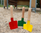Preview: Drei Sandschaufeln für Kinder stecken nebeneinander im Sand eines Spielplatzes. Sie haben einen roten, einen grünen und einen gelben Besatz.